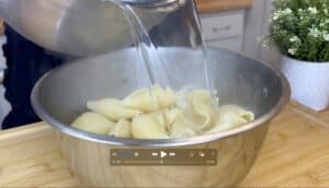 Pouring hot water over pasta shells in a metal bowl on a wooden counter.